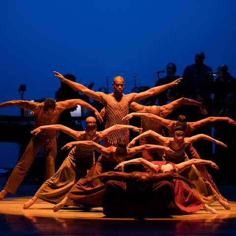 Nine Black dancers are in a cluster formation with arms extended outwards on each side of their body, head and shoulders slightly bent down, one stands upright in the centre, the others all around forming four rows. The dancer in the front row crouches down his head facing the floor. A yellow spotlight on them creates a contrast with the blue background behind them where nine musicians in shadow play instruments.
