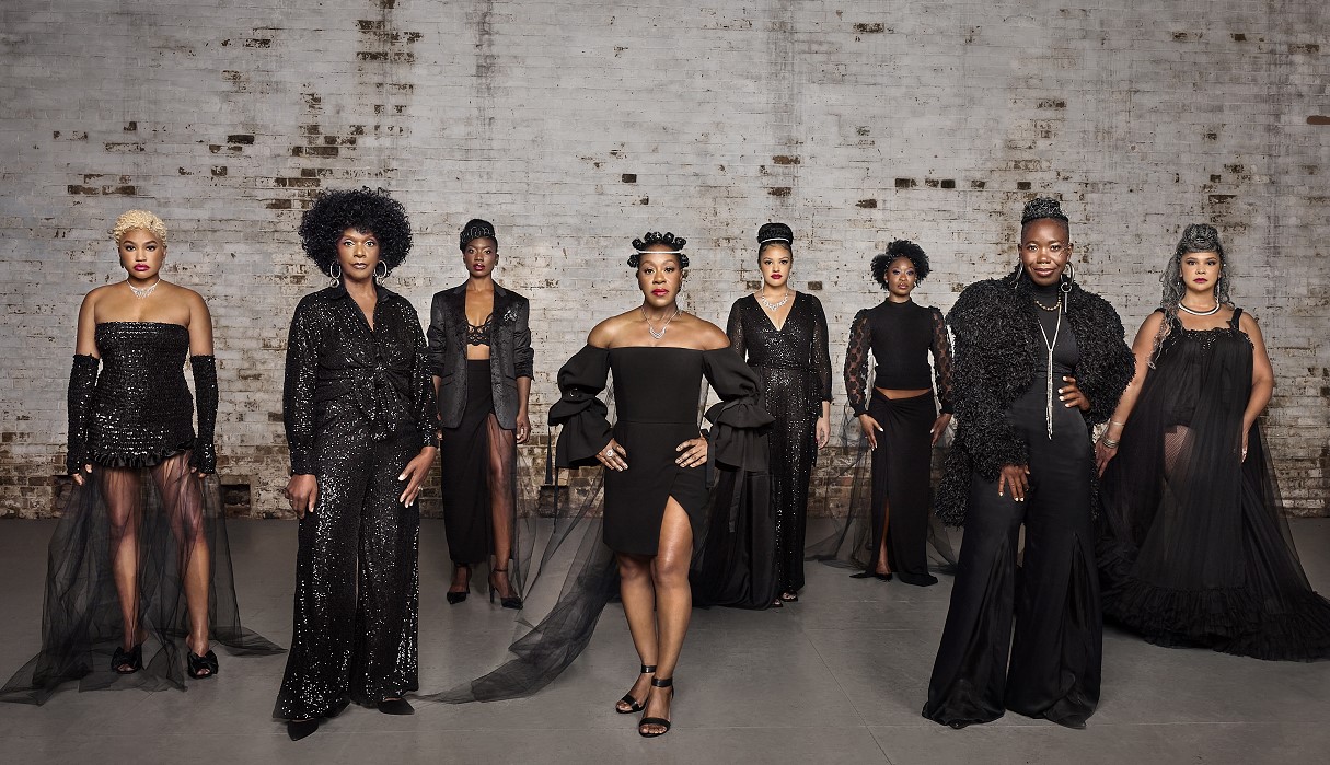 The cast of the show posed in glam black outfits, 8 black women pose in front of a distressed white brick wall. The actesses are Olunike Adeliyi, Chelsea Russell, Ordena Stephens-Thompson, Shakura Dickson, Satori Shakoor, Miranda Edwards, d'bi.young anitafrika and Tamara Brown.