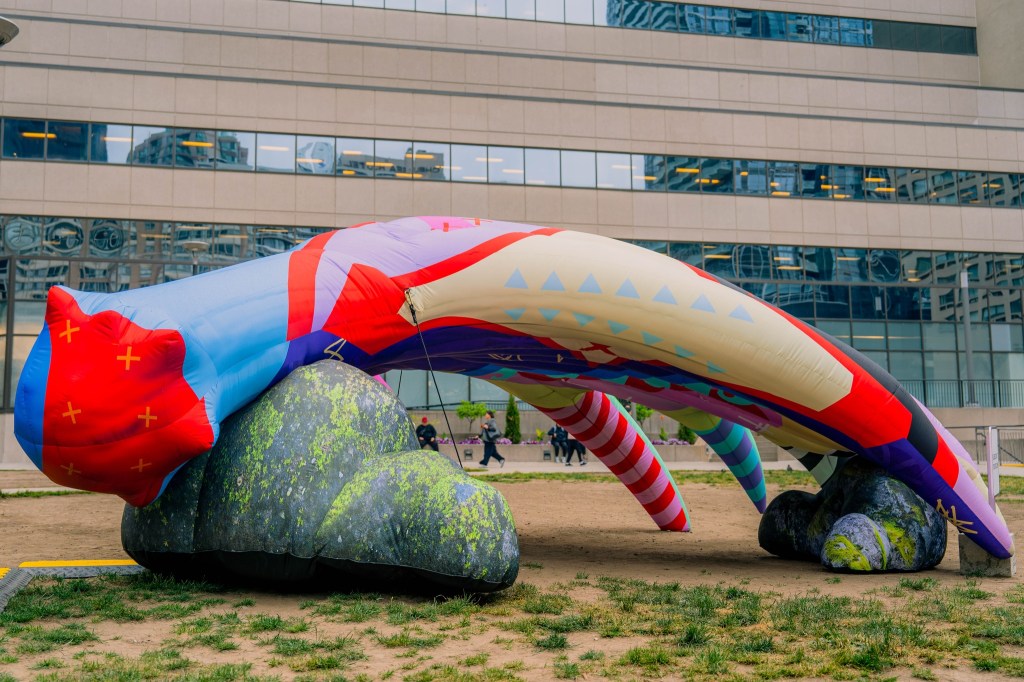 A multicolored work of public art. This giant inflated structure is in the shape of the antler of a moose with bright purple, green, yellow, blue, black, white, red and cyan. It rests on rocks on a grass area in front of building. People walk in the background.